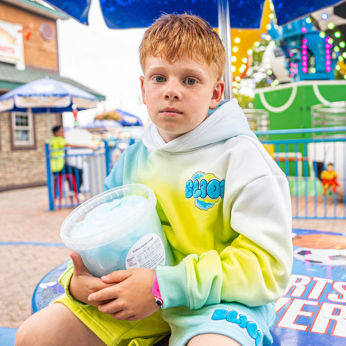 Child in a gradient blue and yellow hoodie holding cotton candy at an amusement park.