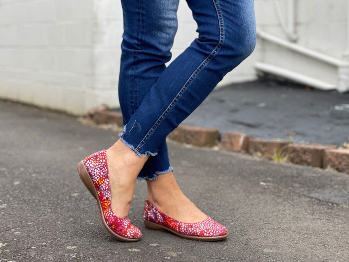 A person wearing blue jeans and red ballet flats with a multicolored design on the shoe's surface. Comfortable European women's shoes.