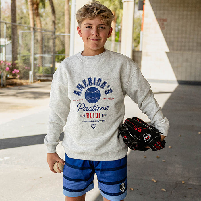 Smiling boy holding a baseball and glove at a baseball facility wearing a gray heather America's Pastime crewneck sweatshirt and royal blue striped mesh shorts