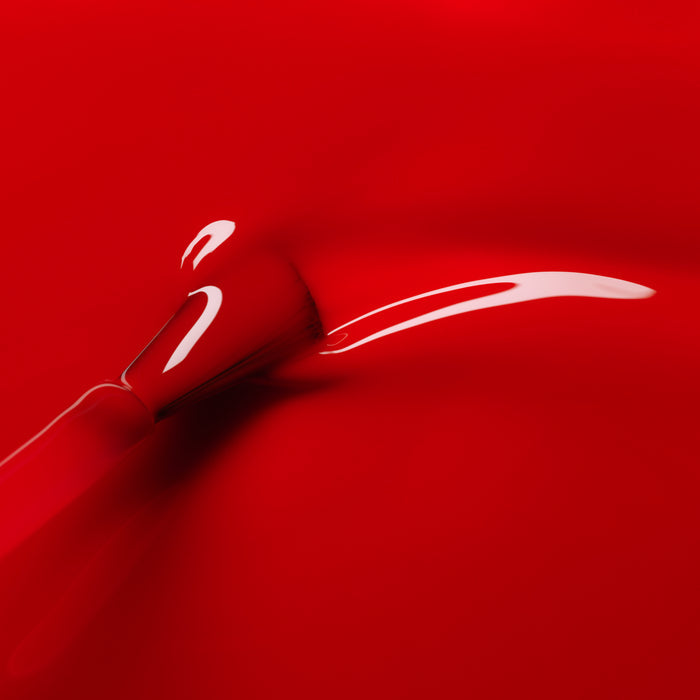 A close-up of a brush applying Anémone by Manucurist, creating a smooth, curved stroke in glossy, vibrant red with luminous reflections on the wet surface.