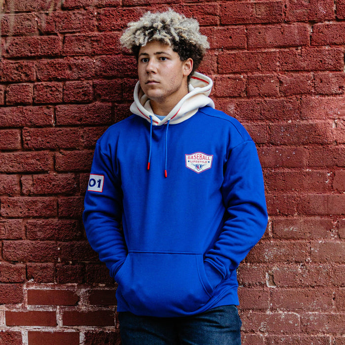 Man with hands in pockets leaning against a brick wall wearing a royal blue champions hoodie with a cream hood
