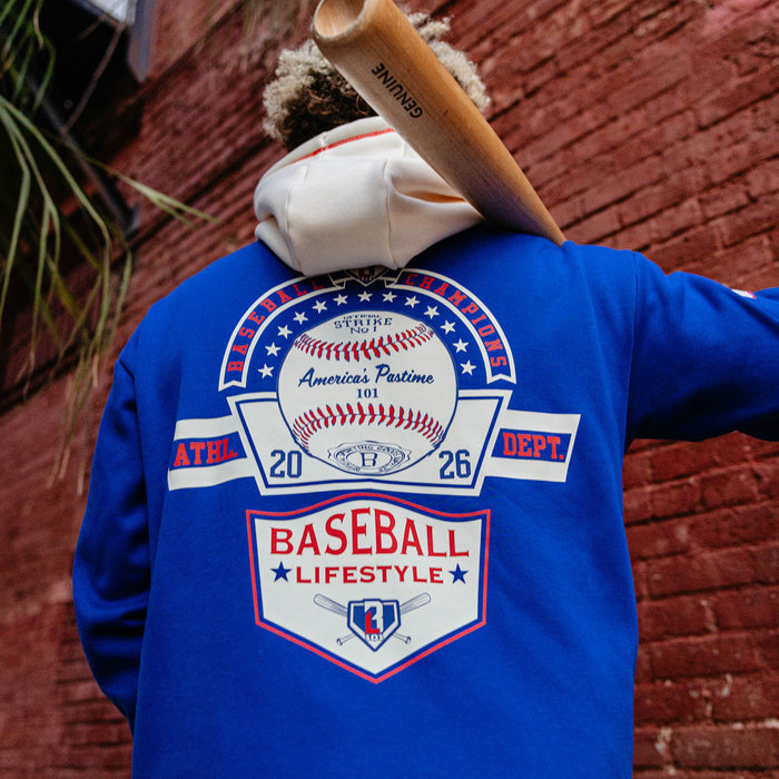 Back view of man holding a bat on his shoulder against a brick wall wearing a royal blue champions hoodie with Baseball Champions America's Pastime back graphic