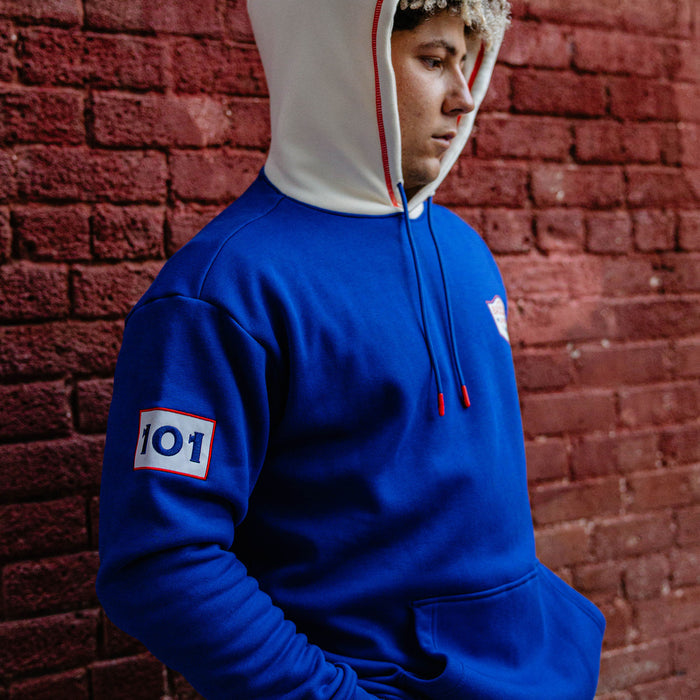 Man looking sideways against a brick wall wearing a royal blue champions hoodie showing the 101 sleeve patch and cream hood