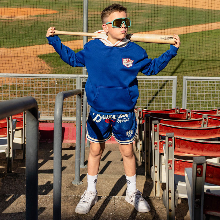 Boy holding a bat across his shoulders at red stadium bleachers wearing teal shield sunglasses and a royal blue champions hoodie with royal blue Baseball Lifestyle mesh shorts