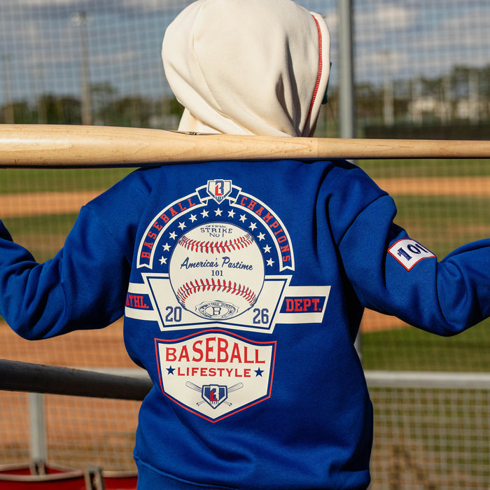 Back view of boy holding a bat across his shoulders at a baseball field wearing a royal blue champions hoodie showing the Baseball Champions America's Pastime back graphic and 101 sleeve patch