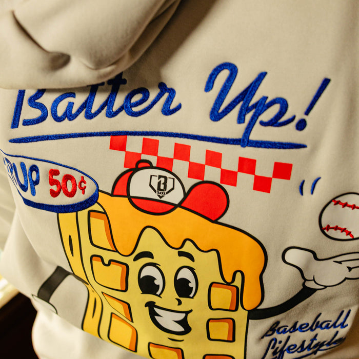 Close-up of a hoodie with a cartoon waffle character and 'Batter Up!' text on a beige background