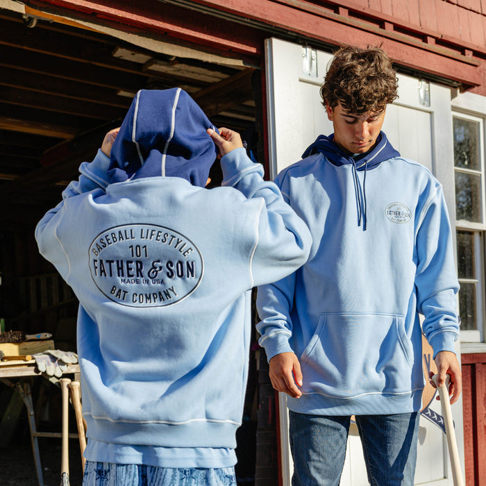 Two male models wearing the columbia blue BL101 Bat Co Hoodie showing both the front chest logo and the large Father & Son Bat Company oval graphic on the back.