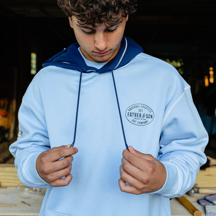 Male model pulling the drawstrings of the columbia blue BL101 Bat Co Hoodie showing the Father & Son Bat Company oval stamp on the chest and navy contrast hood.