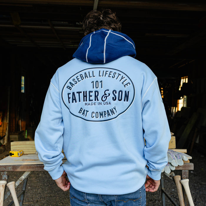 Back view of a male model wearing the columbia blue BL101 Bat Co Hoodie showing the large Father & Son Bat Company oval graphic and navy contrast hood.