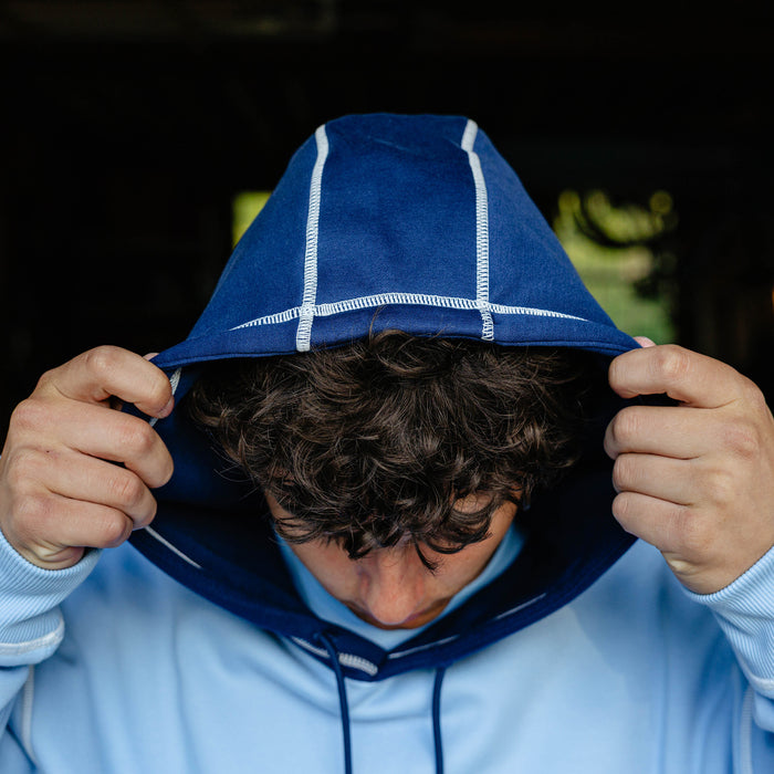 Close-up of a male model pulling the navy contrast hood of the columbia blue BL101 Bat Co Hoodie showing the white contrast stitching detail.