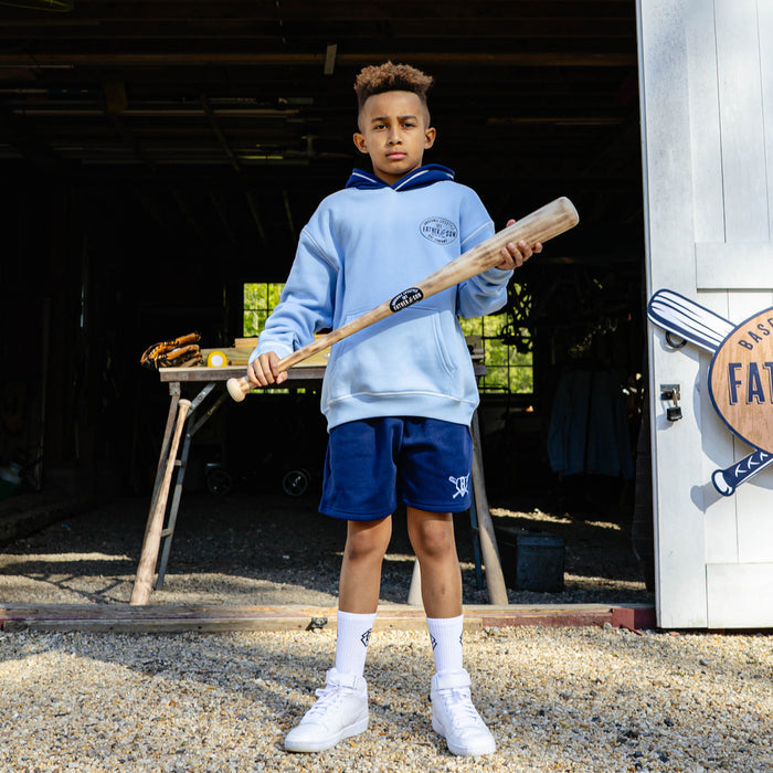 Young boy holding a wooden baseball bat and wearing the columbia blue youth BL101 Bat Co Hoodie with navy contrast hood, paired with navy Heritage Diamond Sweatshorts and white BL101 crew socks.