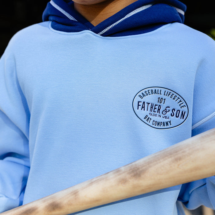 Close-up of the columbia blue youth BL101 Bat Co Hoodie showing the Father & Son Bat Company oval stamp on the chest and navy contrast hood with white topstitching.