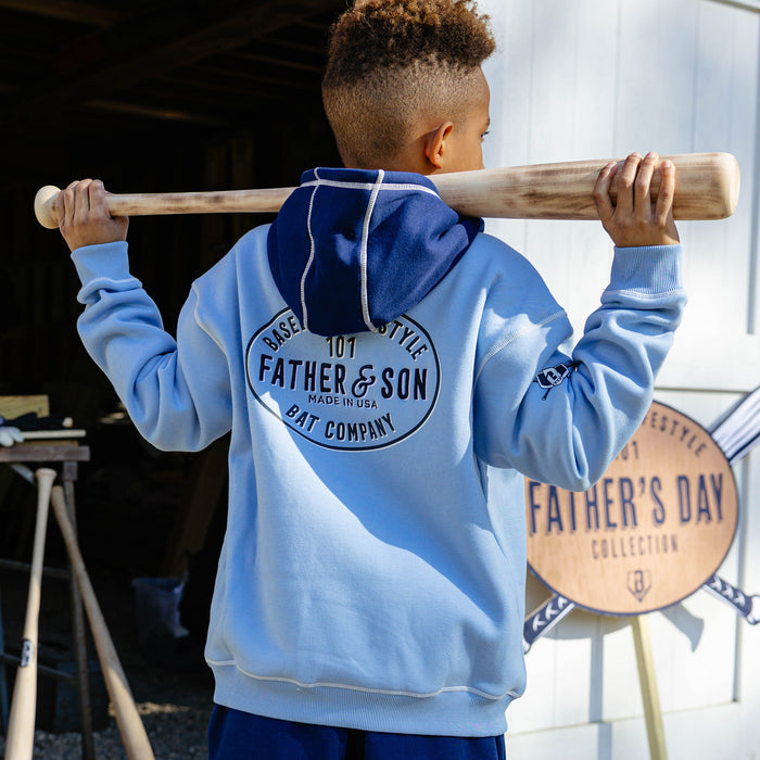 Back view of a young boy holding a baseball bat across his shoulders wearing the columbia blue youth BL101 Bat Co Hoodie showing the large Father & Son Bat Company oval graphic and BL101 sleeve logo.