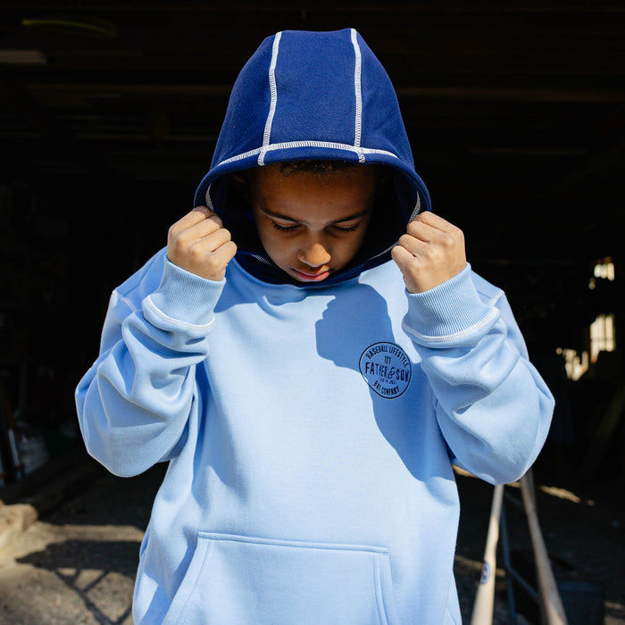 Young boy pulling up the navy contrast hood of the columbia blue youth BL101 Bat Co Hoodie showing the white topstitching detail and Father & Son Bat Company chest stamp.
