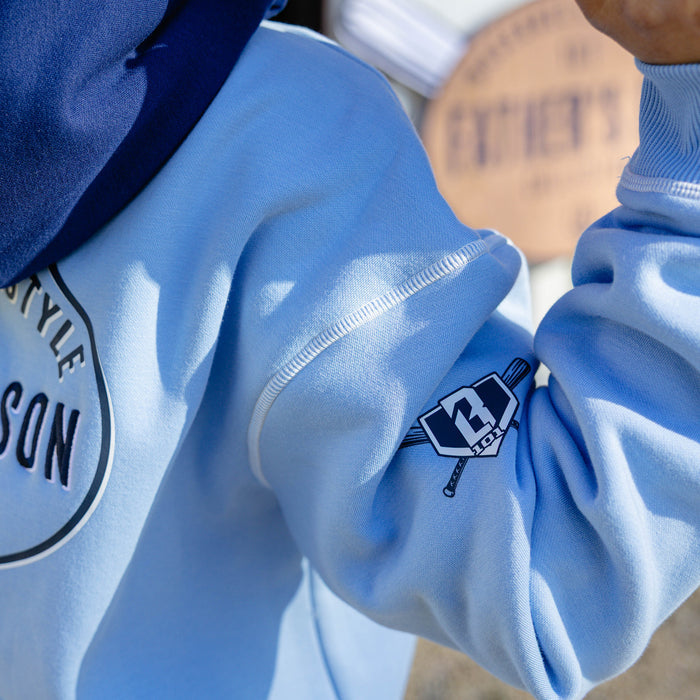Detail shot of the columbia blue youth BL101 Bat Co Hoodie showing the BL101 crossed bats home plate logo on the left sleeve with the Father & Son Bat Company back graphic partially visible.