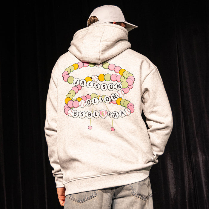 Adult male model seated in a theater holding a baseball glove, wearing the heather gray Jackson Olson x BL101 BSBL Era hoodie with jeans