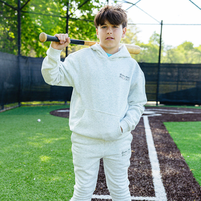 Youth player holding a bat over his shoulder on a baseball field, wearing the heather gray Jackson Olson x BL101 BSBL Era hoodie and matching sweatpants
