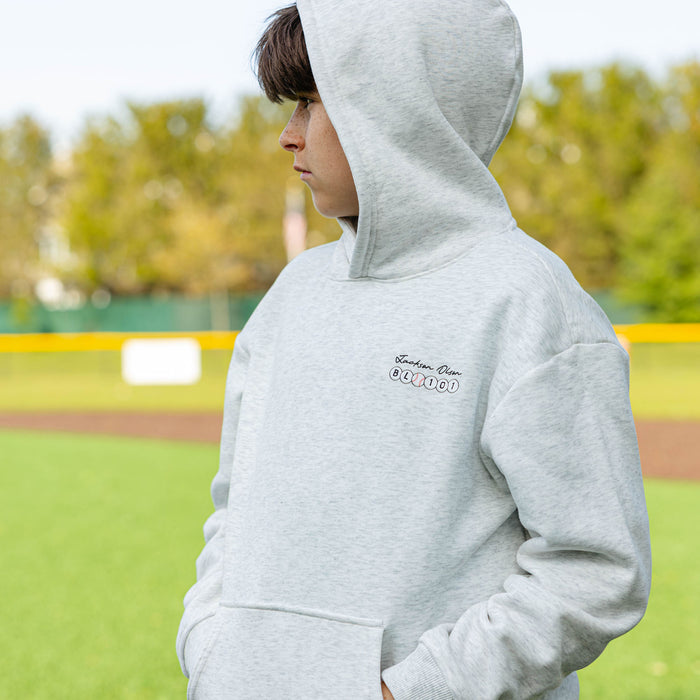 Side profile of a youth player on a baseball field wearing the heather gray BSBL Era hoodie, showing the "Jackson Olson" script and BL101 baseball bead chest logo