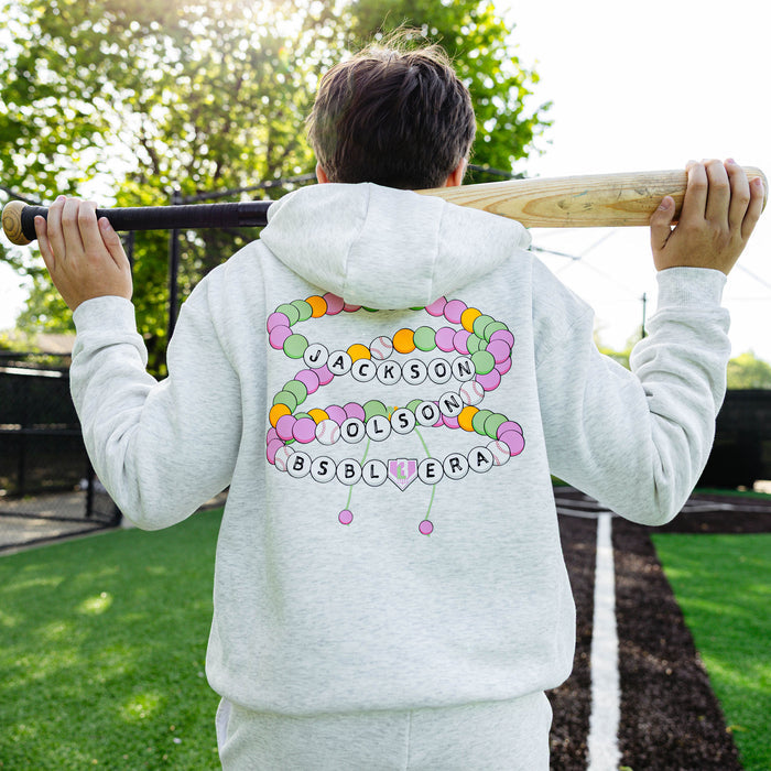 Back view of a youth player holding a bat over his shoulders, showing the colorful friendship bracelet graphic with "Jackson Olson BSBL Era" lettering on the heather gray BSBL Era hoodie