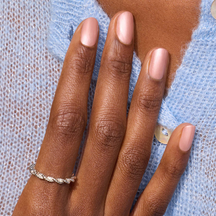 A hand with Manucurist’s Carnation pearly pink beige nail polish rests on a light blue knit sweater, adorned with a twisted silver ring.