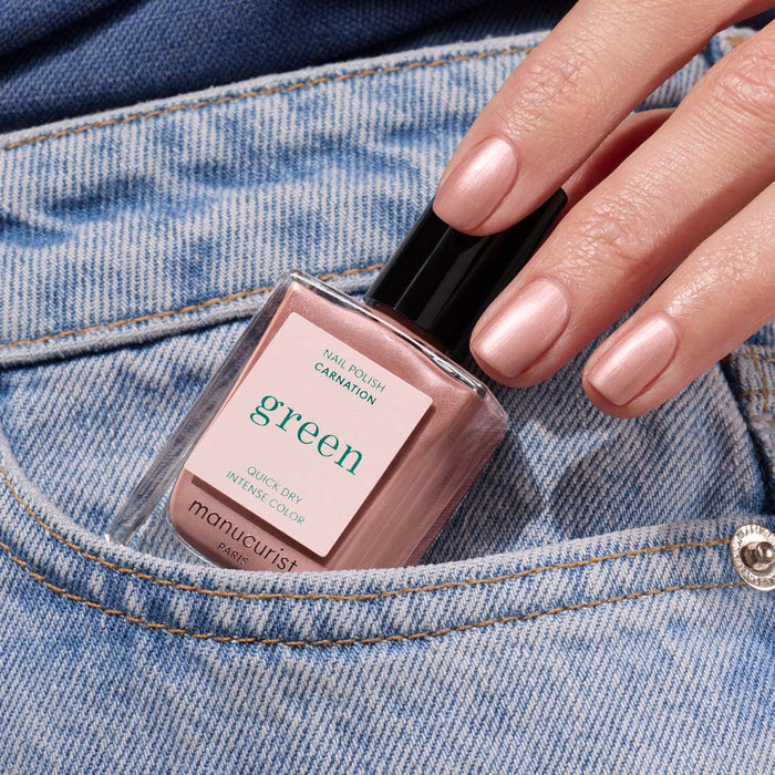 A hand with light pink, glossy shimmer nail polish rests on blue jeans, holding a bottle of Manucurist Carnation nail polish.