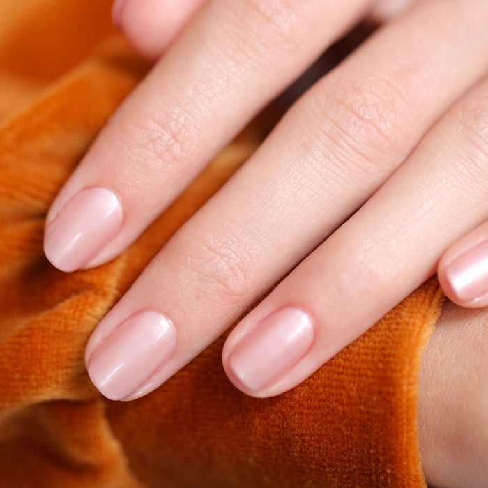 A close-up of a hand with neatly manicured nails painted in Manucurist's Carnation, a pearly pink beige polish, resting on soft orange textured fabric.