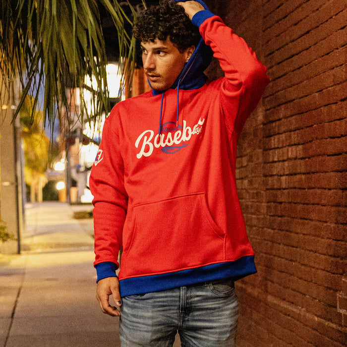 Man leaning against a brick wall at night wearing a red Baseball Lifestyle hoodie with a royal blue hood and jeans