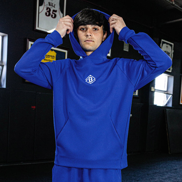 Person wearing a blue hoodie with a logo, standing in a room with sports memorabilia on the wall.