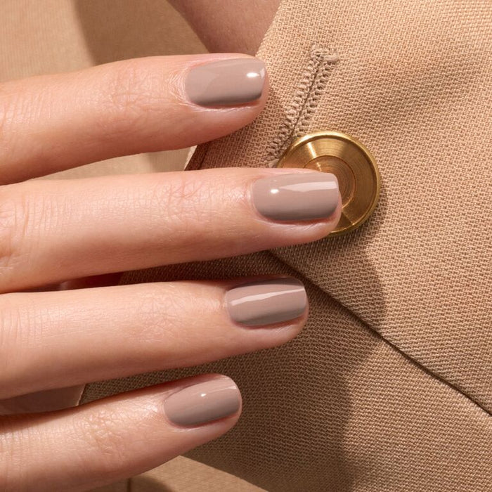 A hand with short, neatly manicured nails painted in Manucurist’s Dove Beige polish rests against a beige fabric with a gold button.