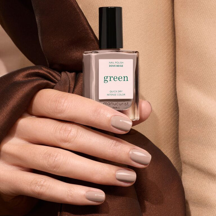 A hand with neatly manicured nails painted in Manucurist Dove Beige holds the matching polish bottle. Soft beige and brown fabrics are in the background.