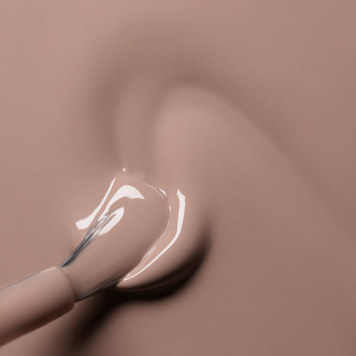 A close-up of a nail polish brush spreading Manucurist’s Dove Beige from the Green™ range on a smooth surface, forming a glossy pastel swirl.