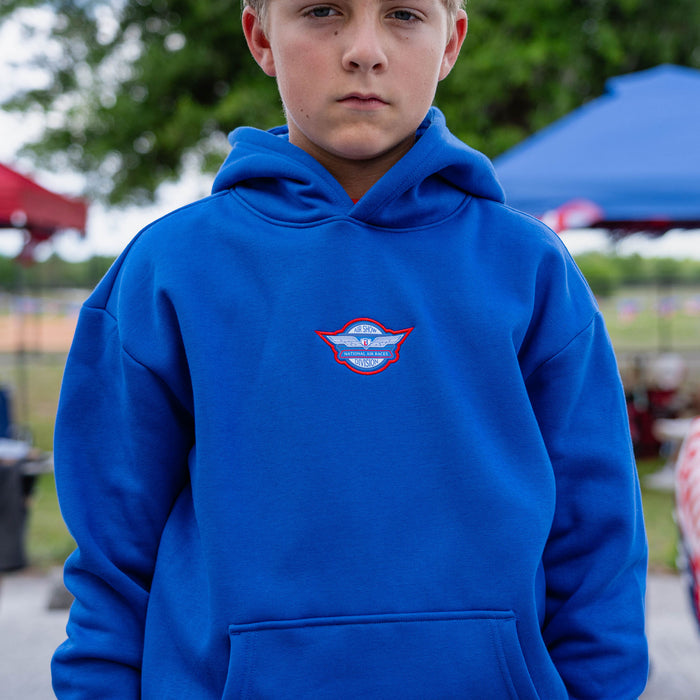 Close-up of youth male model in royal blue Fastball Flight hoodie showing Air Show Division badge on center chest at outdoor event