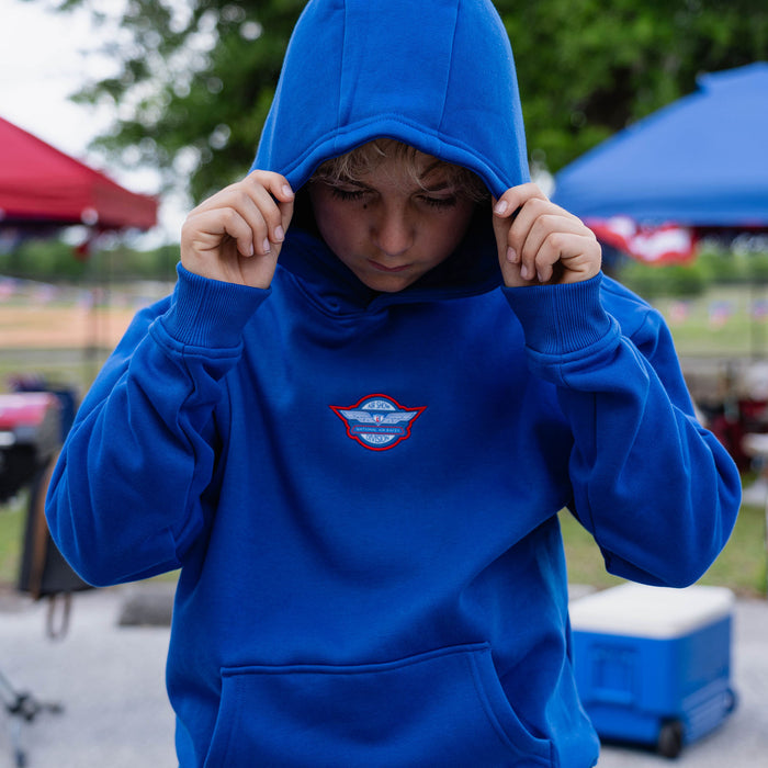 Youth male model pulling hood over head on royal blue Fastball Flight hoodie showing Air Show Division badge on chest at outdoor baseball event