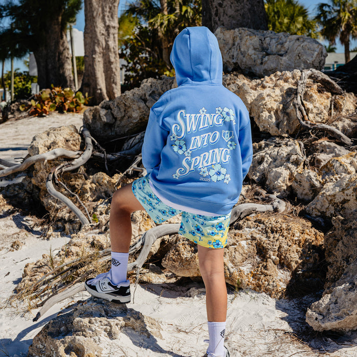 Person wearing a blue hoodie with 'Swing into Spring' text and floral graphics, standing on rocks with palm trees in the background.