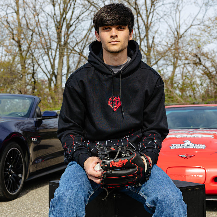 Male model seated outdoors holding a baseball glove and ball wearing a black hoodie with red and white stitched flame detailing on the sleeves and a sports car in the background