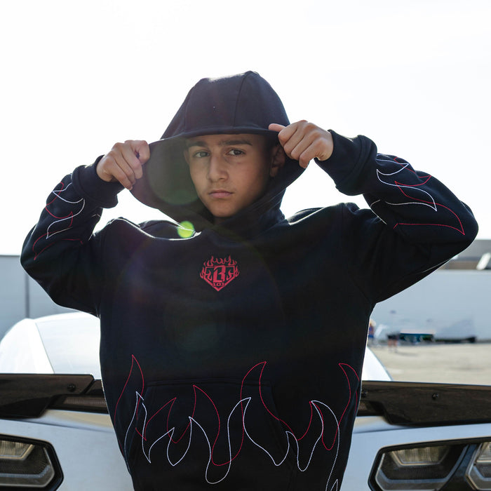 Young male model pulling up the hood of a black hoodie with BL101 flame crest embroidery and red and white stitched flame outlines along the hem and sleeves with a car in the background