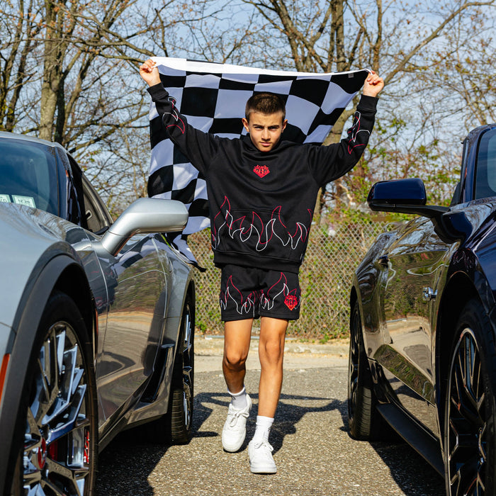Young male model running between two parked cars while holding a checkered flag overhead wearing a black hoodie with flame stitching and matching black shorts