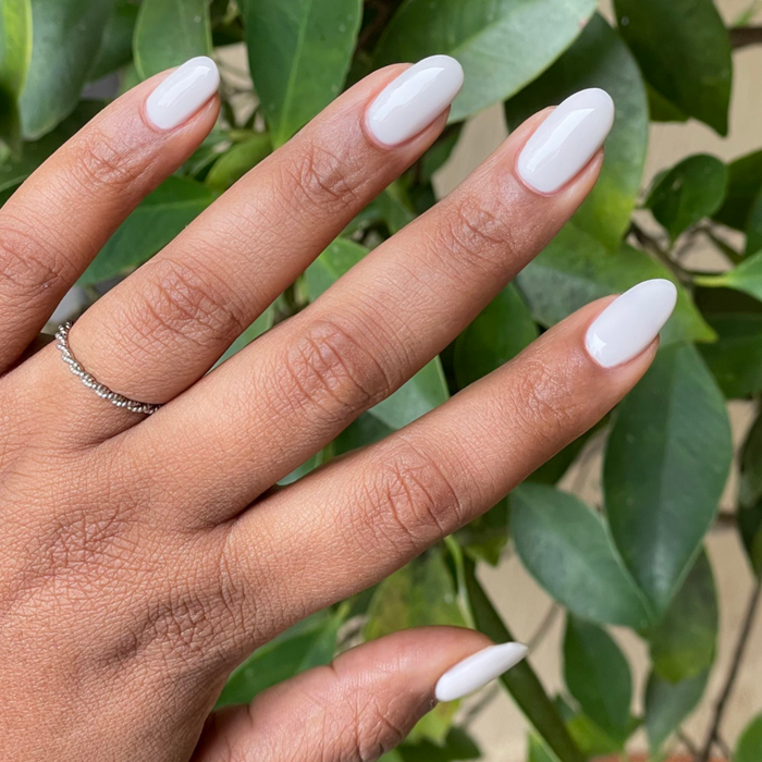 Hand with almond-shaped nails painted in glossy soft white using LHG - Gel polish against leafy green background