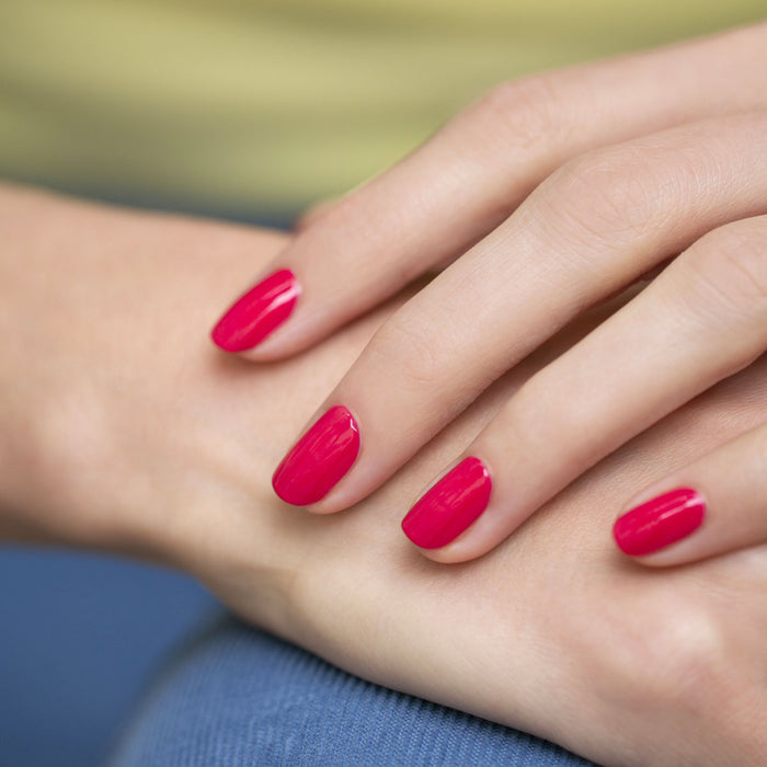 A close-up of a hand with neatly manicured nails painted in Péonie by Manucurist. The fingers rest gently on a surface, and the background is softly blurred in blue and yellow tones.