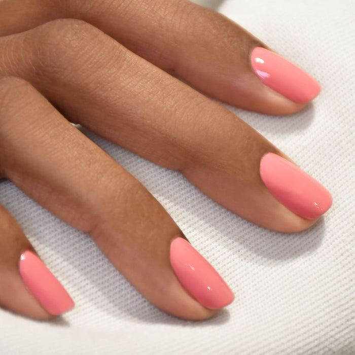 A close-up of a hand with medium brown skin displays neatly manicured nails painted in Pink Paradise by Manucurist, a glossy candy pink shade, resting on white textured fabric.