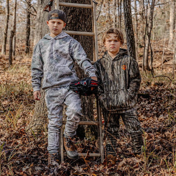 Two children in camouflage clothing standing in a forest with a ladder.
