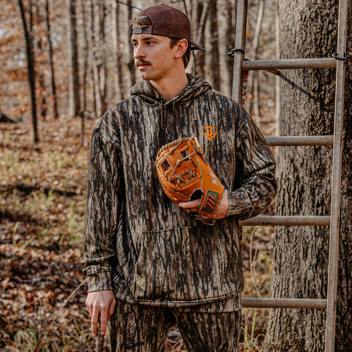 Man in camouflage hoodie and sweatpants, holding a hunting glove in a forest setting