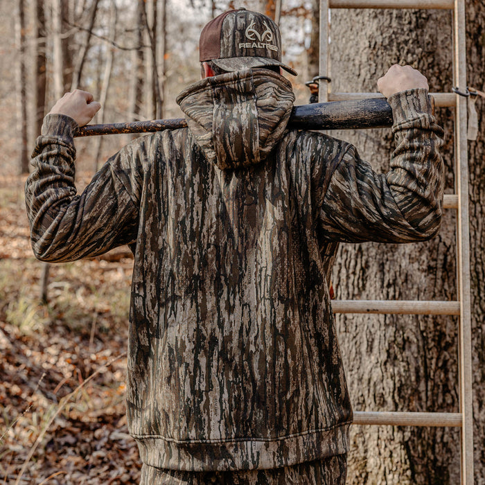 Person in full camouflage gear with a. wooden bat, standing in a forest setting.