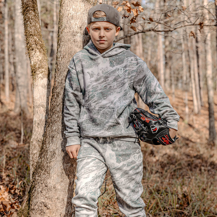 Child in camouflage hoodie and pants holding a baseball glove in a forest setting