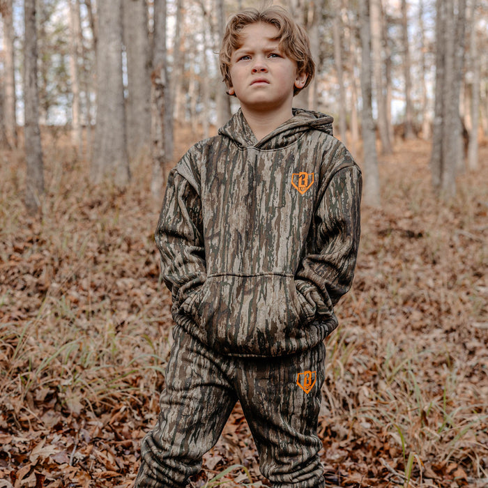 Child wearing a camouflage hoodie and pants with orange BL101 logo standing in a forest.