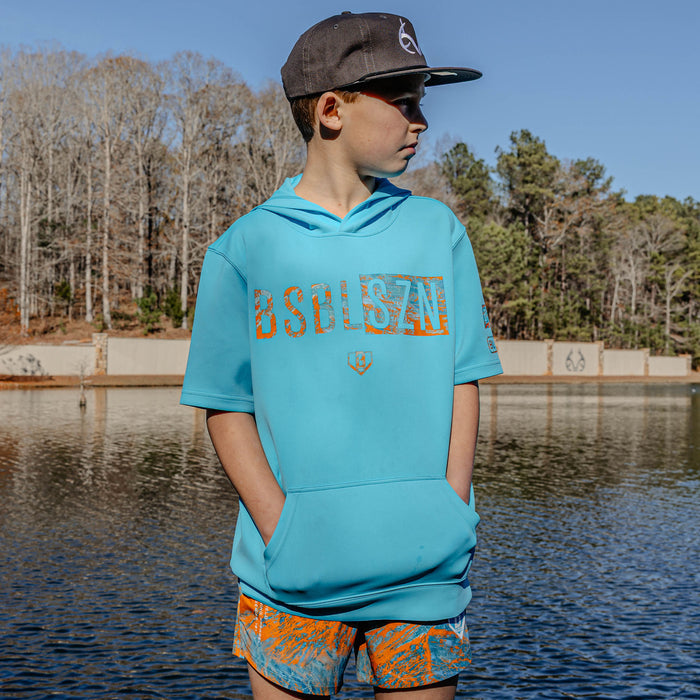 Child wearing a blue hoodie with 'BSBLSZN' text and orange shorts with a lake and trees in the background
