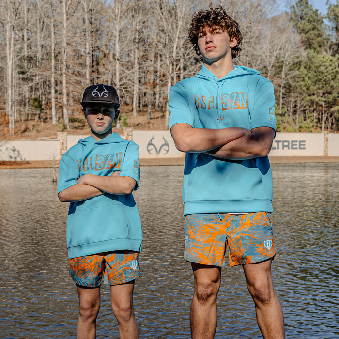 Two individuals wearing matching blue short sleeve hoodies with 'BSBL SZN' logo and orange patterned shorts standing by a body of water.