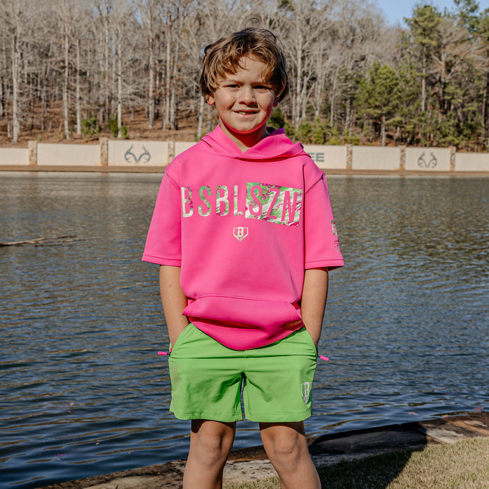 Child wearing a pink hoodie with 'BSBL SZN' text and green shorts standing by a lake with trees in the background