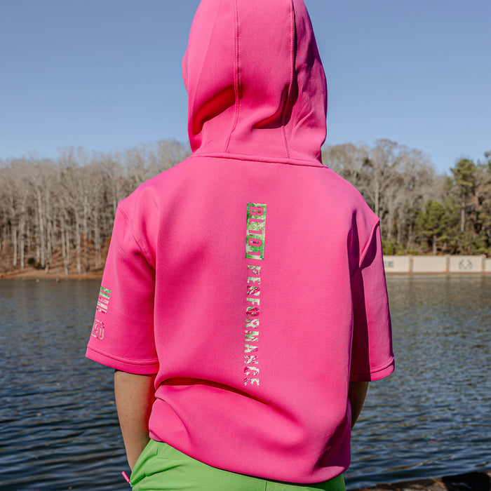 Person wearing a pink hoodie with 'BL101 PERFORMANCE' text on the back, standing by a body of water with trees in the background.