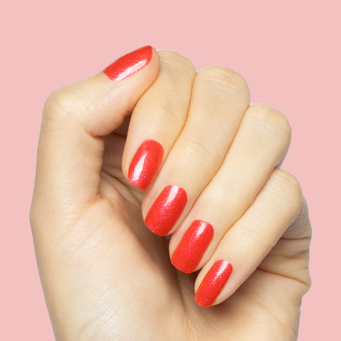 Hand with medium-length nails painted bright pinky-red with silver shimmer Rodeo Queen - Gel polish on soft pink background